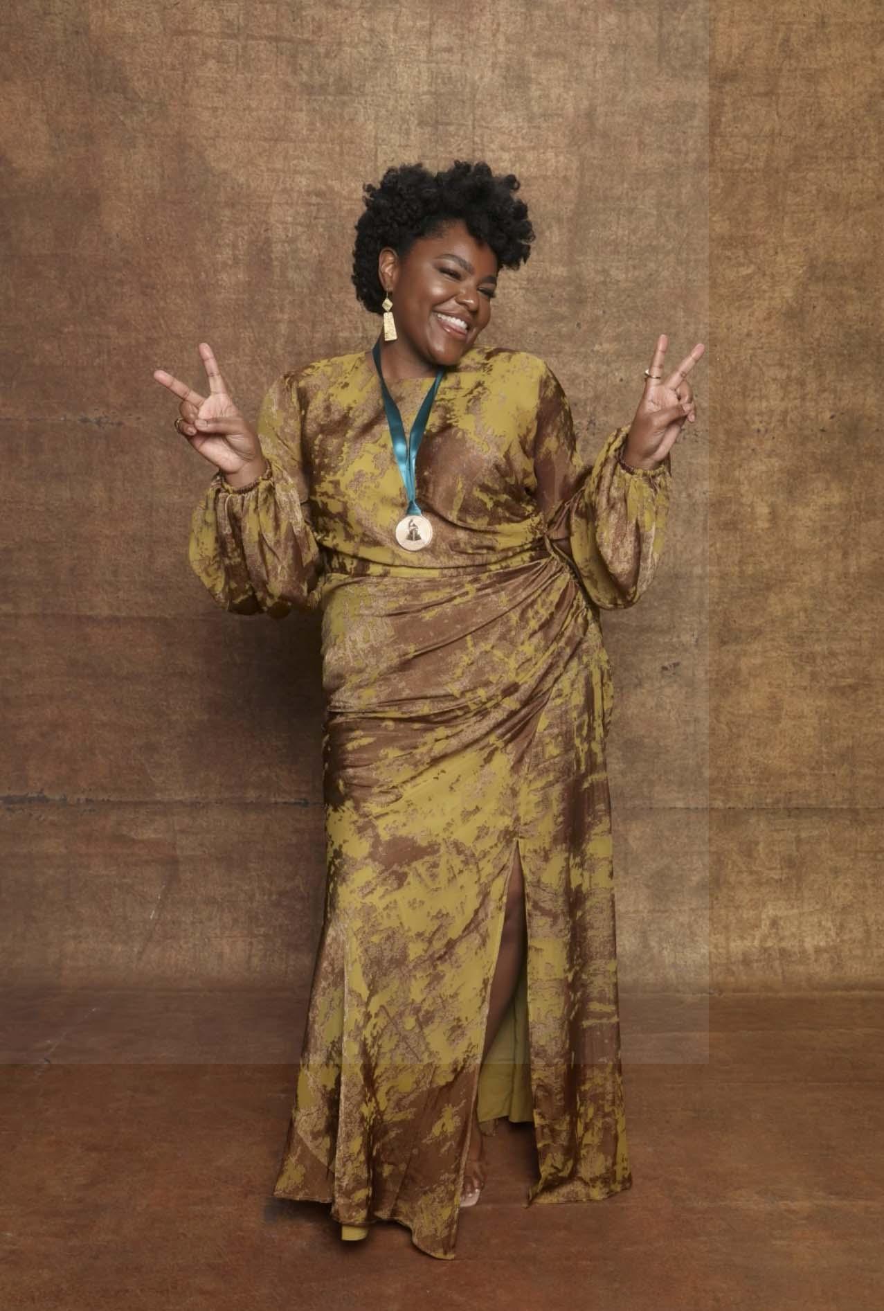 A woman dressed in a golden colored dress. she is holding up the peace sign with both hands