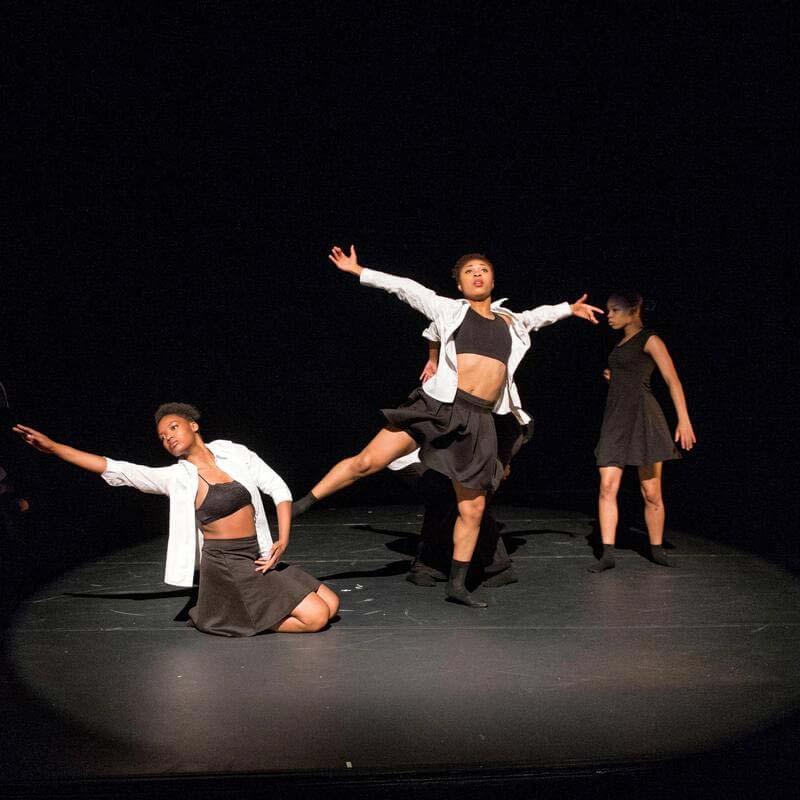 Howard University theatre performers on a dark stage. The dancers are illuminated and they are in different poses. One person is on the floor with their left hand out. the other person in the middle is in mid leap with their hands out