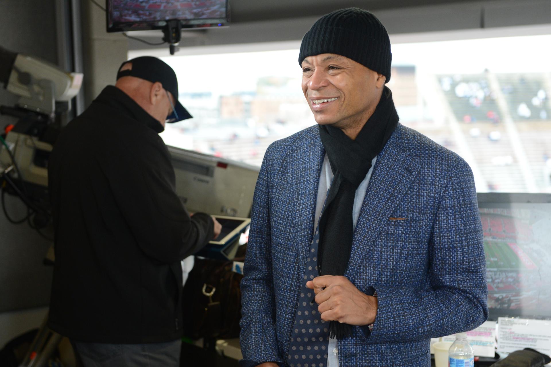 Gus Johnson in a black hat and scarf standing in sports booth