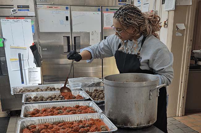 Alternative Spring Break student working in a kitchen. There is a large pot in front as well as pans of food line up in front of her